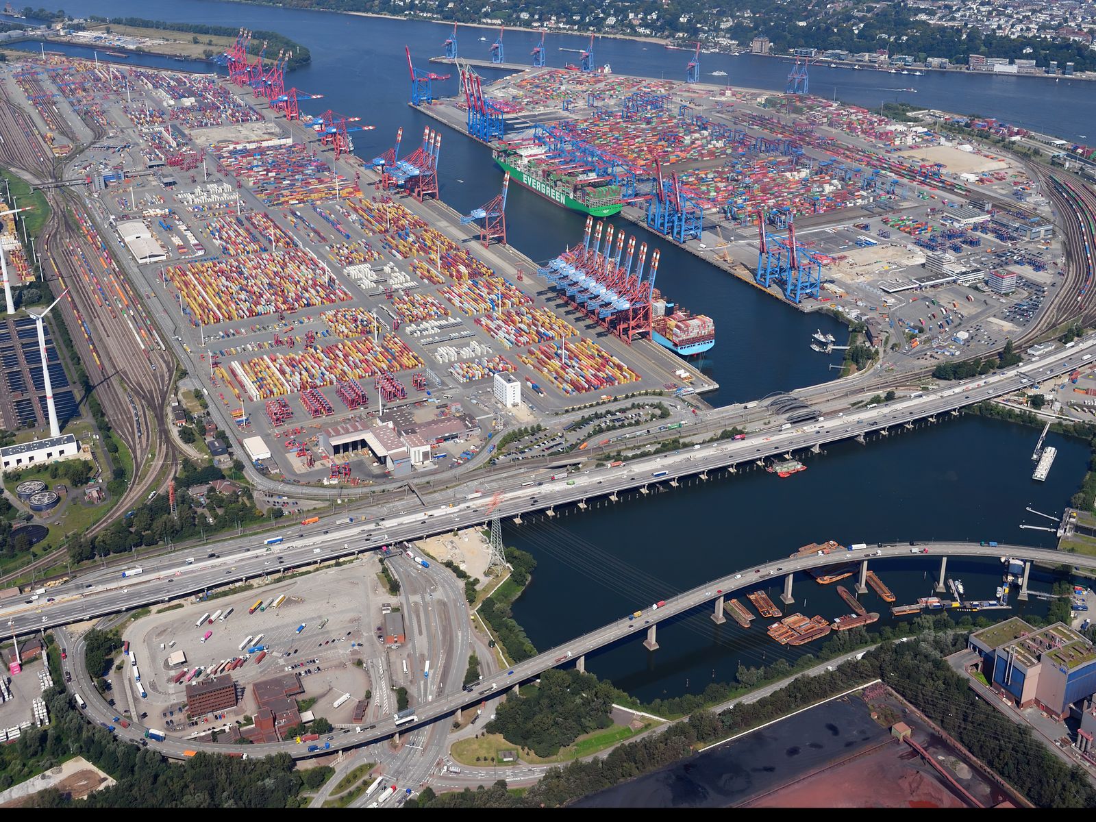 A container terminal in Hamburg from the air: rows of container stacks, gantry cranes and a ship at the quay, the river and a road bridge in front