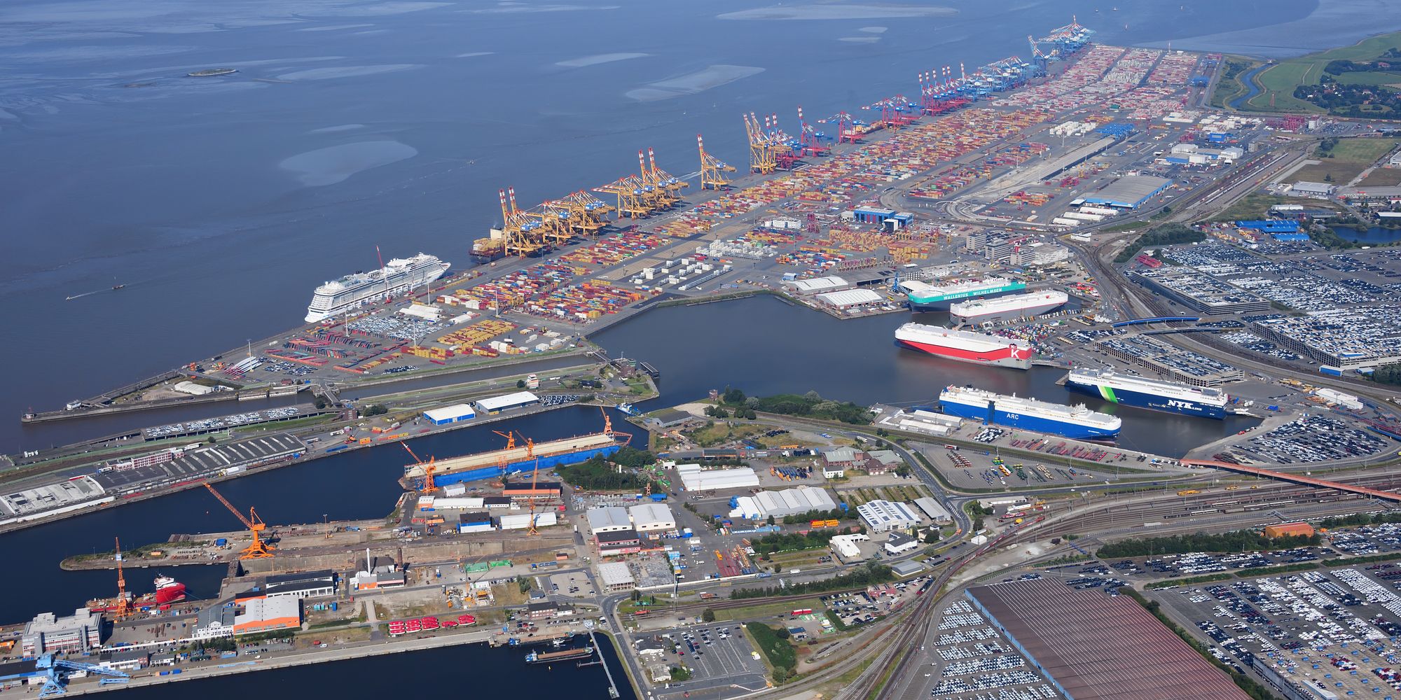 The Bremerhaven container terminal from the air: a long quay of cranes and container stacks along the sea, car carriers in the harbour basin
