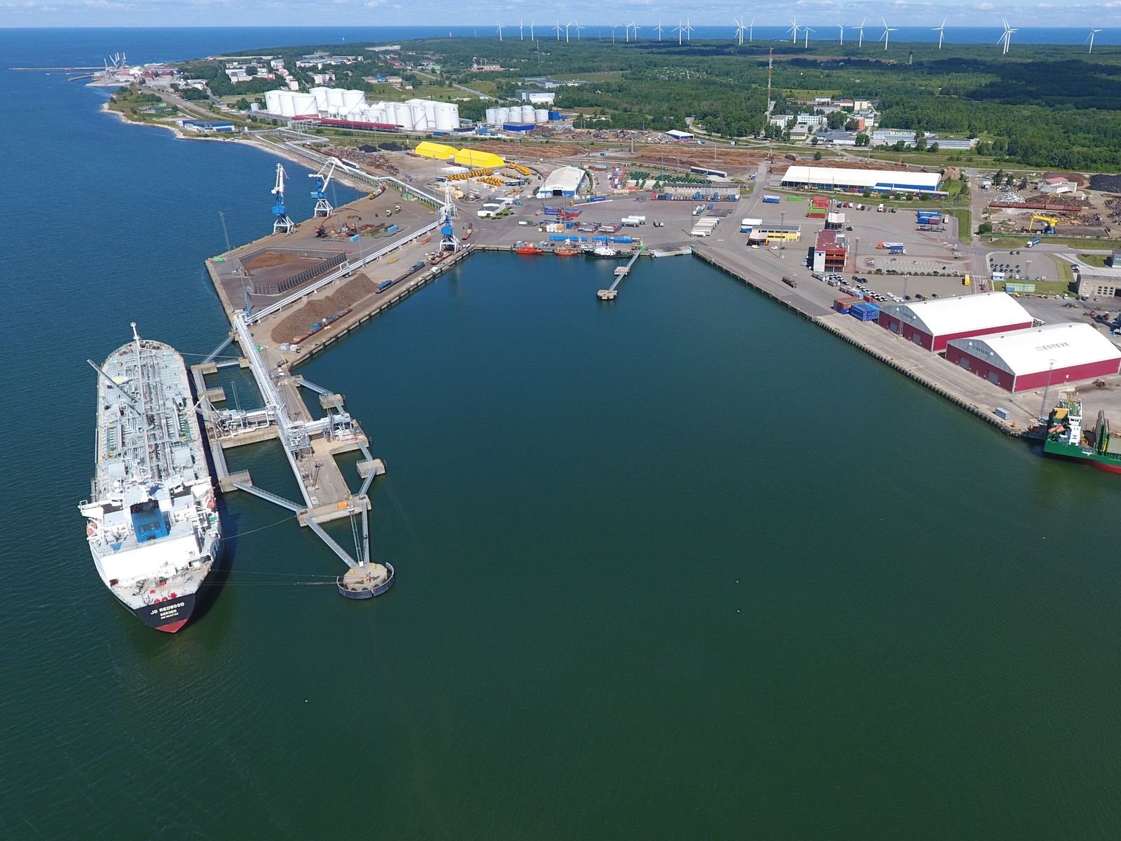 Paldiski South Harbour from the air: a tanker at the jetty, quays, warehouses and storage tanks, wind turbines on the horizon