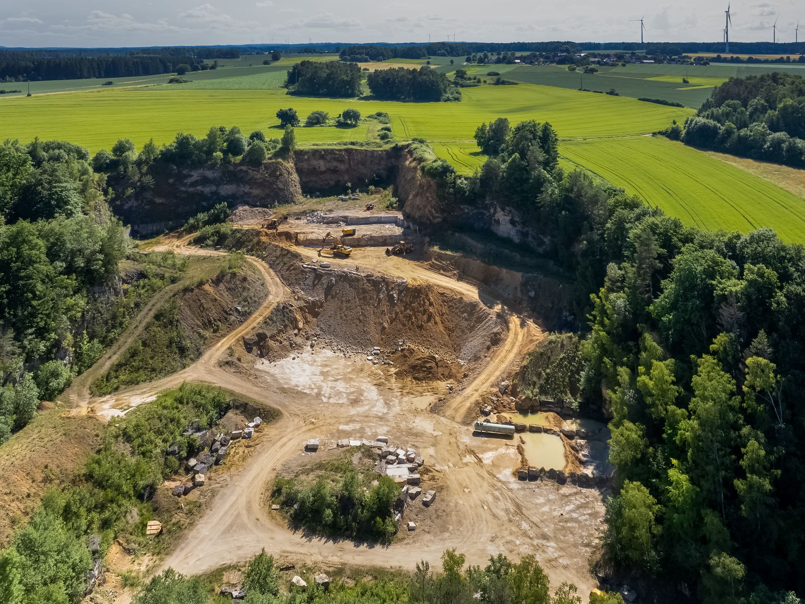 A working limestone quarry from the air: haul roads winding down to the pit floor, diggers and cut blocks, forest and fields around