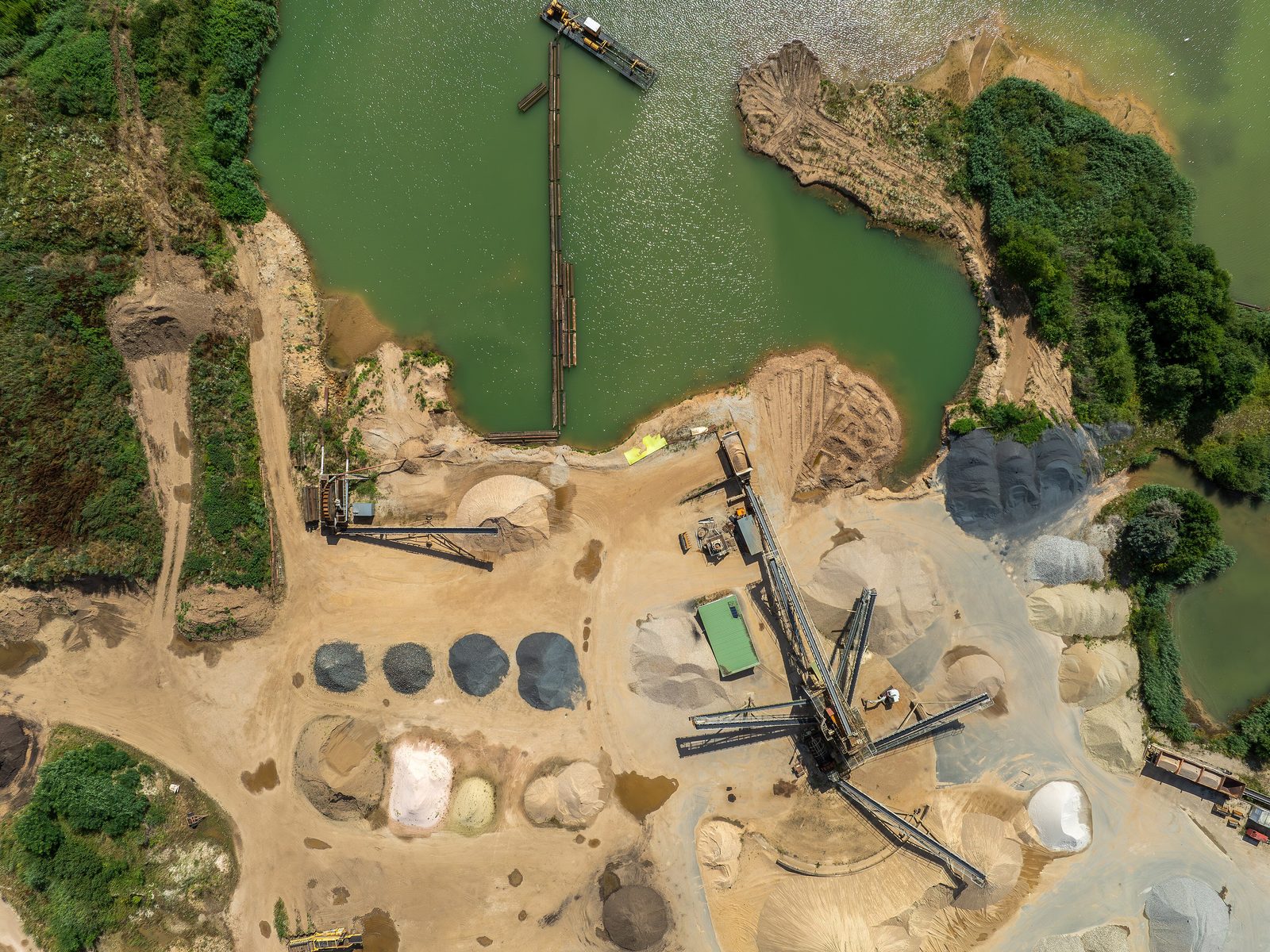A gravel pit from straight above: cone-shaped piles of sand and gravel, a conveyor and a dredger on a green lake