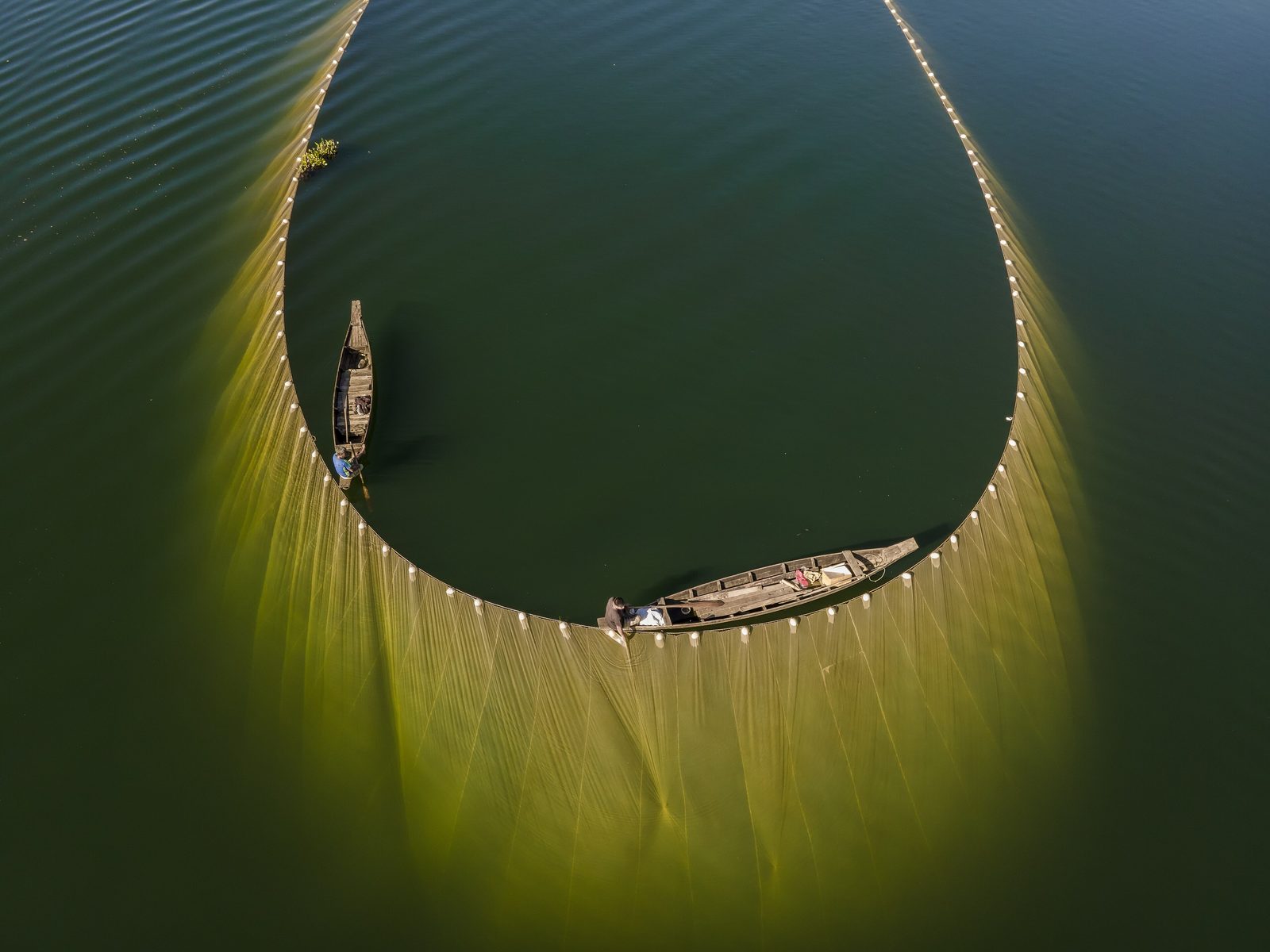A fishing net from straight above on a calm lake: the floats in a long loop and the net hanging down into the water, two small boats inside it