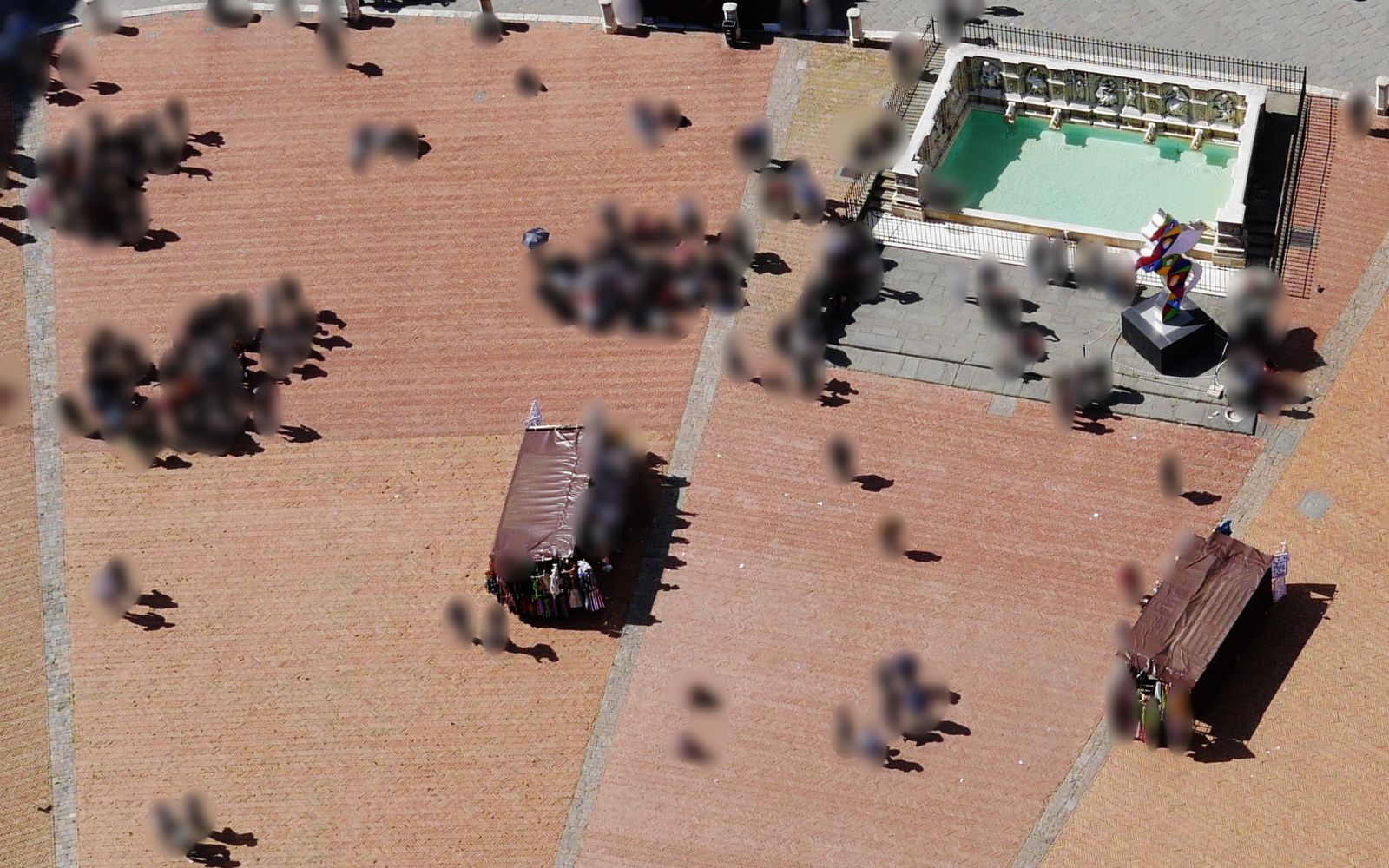 The same square with every person blurred into a soft smudge; the fountain, the stalls and the shadows stay sharp
