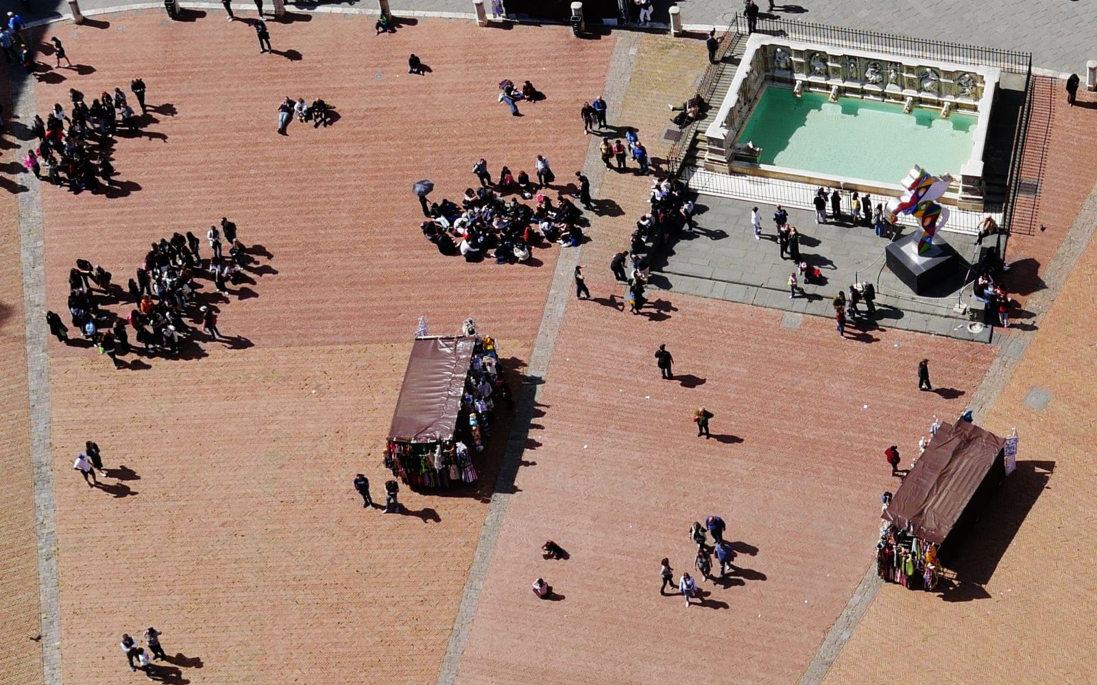 Piazza del Campo in Siena from high above: groups of people walking and sitting on the brick square, a fountain and two market stalls