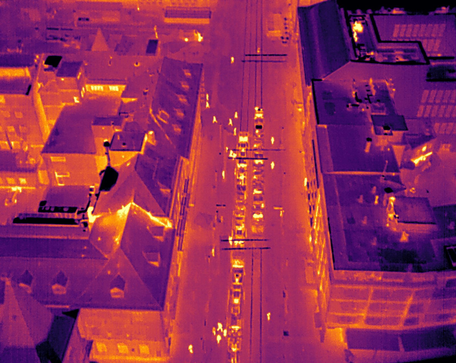 A thermal picture of roofs and a street from a drone: the warm street and roof edges glow orange in the dark
