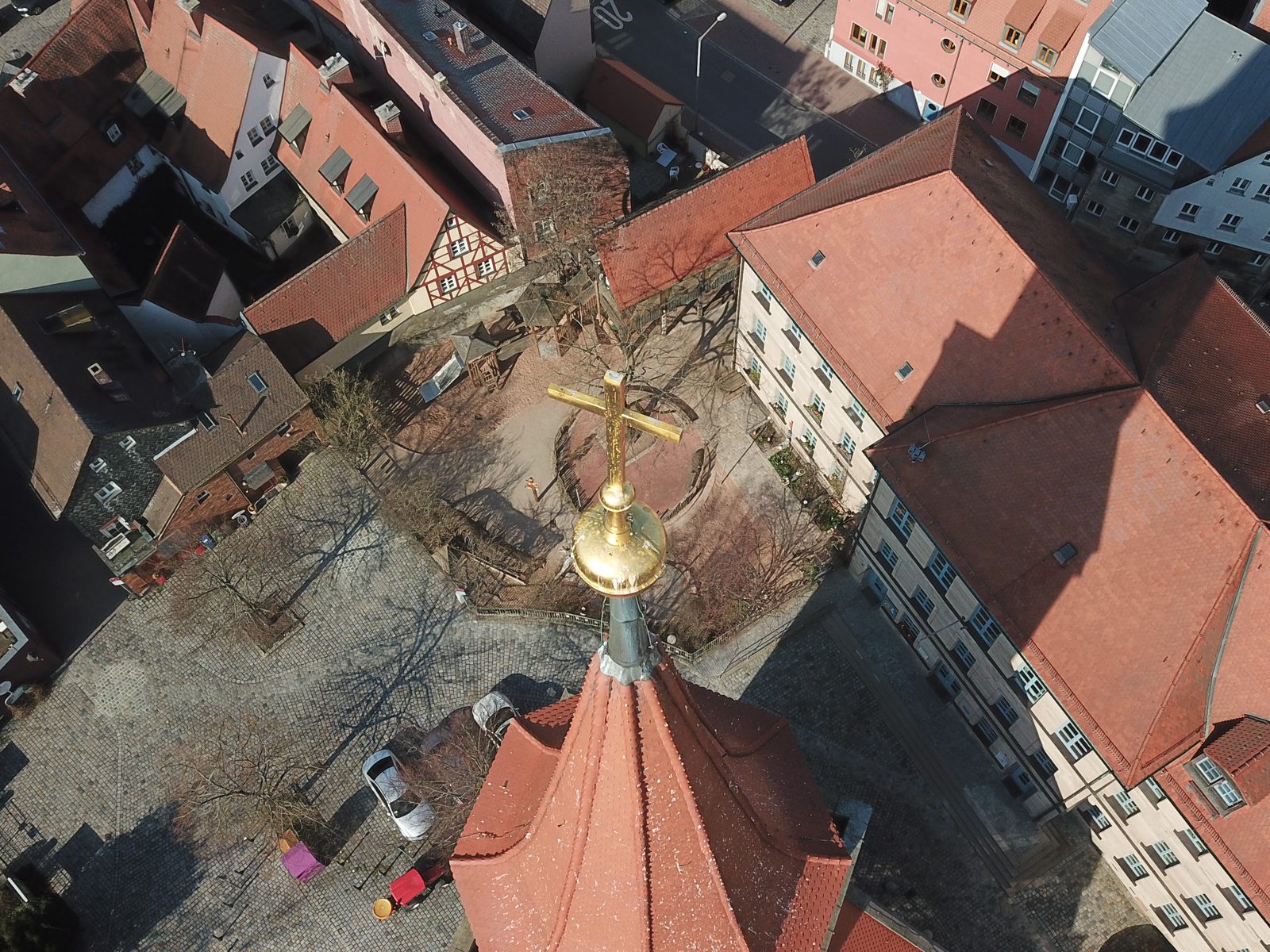 A church spire with a golden cross seen from a drone above it, the old town square far below